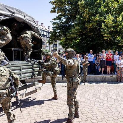 Zdjęcie przedstawia żołnierzy w kamuflażu i sprzęcie taktycznym wysadzających się z wojskowego pojazdu przed publicznością. Szkolenie związane z ewakuacją w sytuacjach kryzysowych. (wygenerowano przez AI)