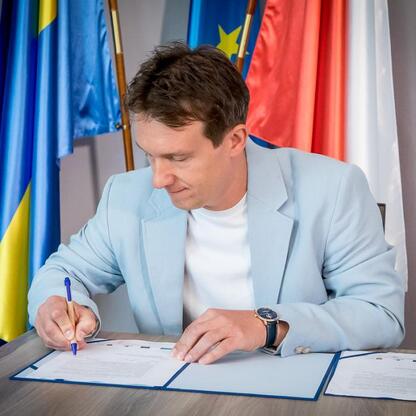 Mężczyzna w niebieskim garniturze podpisuje dokumenty, siedząc przy biurku w formalnym pomieszczeniu z flagami Europy. (wygenerowano przez AI)