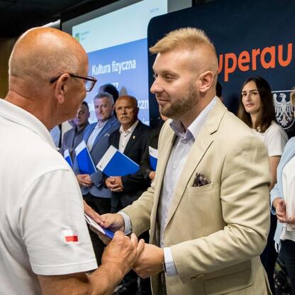 Na ceremonii w Stadionie Śląskim, organizowanej przez Superauto.pl, dwaj uczestnicy w szarej i białej koszulce trzymają się za ręce, tło z polskim emblematem i napisem 