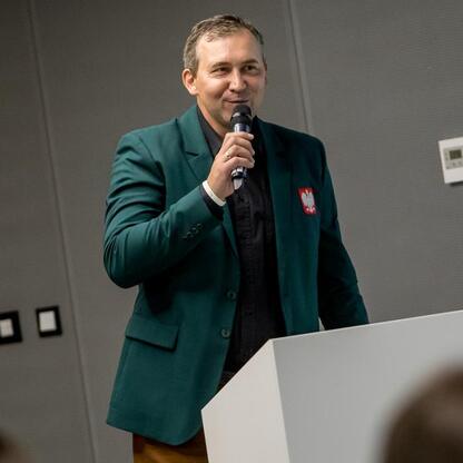 Na podium w zielonym garniturze z orłem, mówiąc do publiczności. (wygenerowano przez AI)