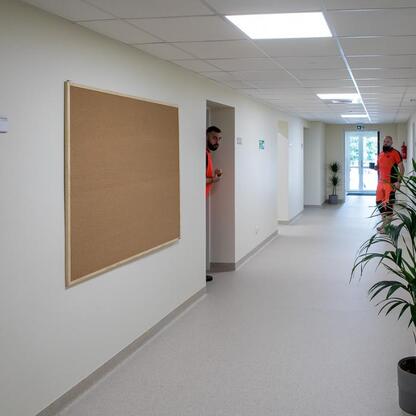 Modernizowana korytarz w siedzibie Rudzkiego Pogotowia Ratunkowego. Oświetlony sufitem, z białymi ścianami, korkoboardami i roślinami. Dwaj pracownicy w pomarańczowych mundurach. (wygenerowano przez AI)