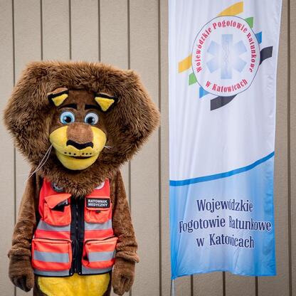 Mascot dressed as a brown bear in a red medical vest stands in front of a banner for Wojewódzkie Pogotowie Ratunkowe in Katowice. (wygenerowano przez AI)