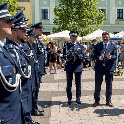 Formalna uroczystość na rybnickim rynku, gdzie obchodzono Święto Policji w 2023 roku. Uniformowani policjanci i urzędnicy w garniturach gestykulują, tło to tłoczna ulica z flagą Polski. (wygenerowano przez AI)