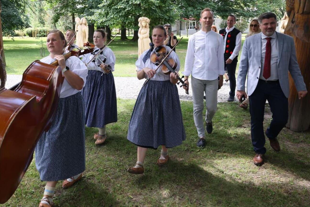 Fotografia przedstawia moment z Tydzień Kultury Beskidzkiej, gdzie kobieta w białej bluzie i czerwonym naszyjniku rozmawia z dziennikarzem, używając mikrofonu. Scena rozgrywa się na zewnątrz w parku. (wygenerowano przez AI)