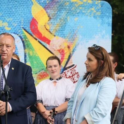 Festiwal Kultury Beskidzkiej: Mężczyzna w garniturze przemawia do mikrofonu, kobieta w niebieskim płaszczu stoi obok, na tle mosaiku i muzyków grających na instrumentach strunowych. (wygenerowano przez AI)