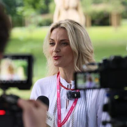 Fotografia przedstawia moment z Tydzień Kultury Beskidzkiej, gdzie kobieta w białej bluzie i czerwonym naszyjniku rozmawia z dziennikarzem, używając mikrofonu. Scena rozgrywa się na zewnątrz w parku. (wygenerowano przez AI)