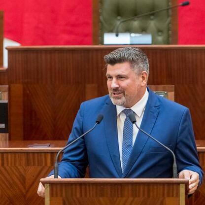 Mężczyzna w niebieskim garniturze przemawia na podium w formalnym uroczystości, związany z dostarczeniem nowych wozów strażackich dla OSP w regionie Śląskim. (wygenerowano przez AI)