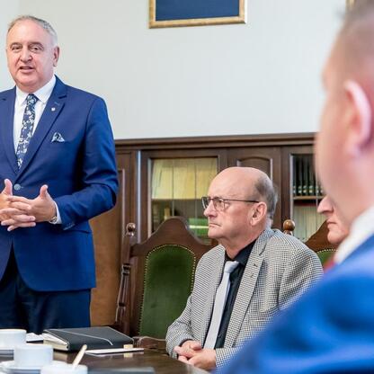 Formalna sesja w sali konferencyjnej, gdzie mężczyzna w niebieskim garniturze prezentuje dokumenty, podczas gdy grupa ludzi słucha uważnie. (wygenerowano przez AI)