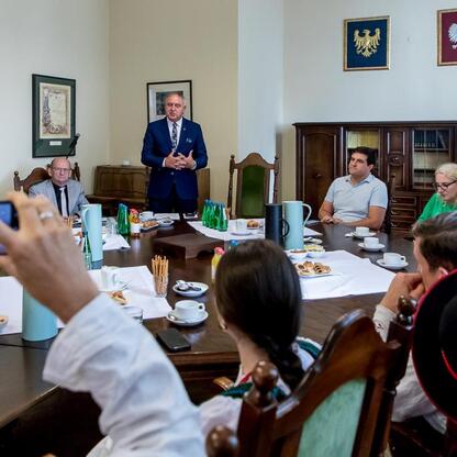 Formalna sesja wojewódzkiego programu Owca Plus w budynku administracyjnym, gdzie menedżer w garniturze przemawia do grupy ludzi, podkreślając znaczenie ochrony przyrody i krajobrazu poprzez wypas owi (wygenerowano przez AI)