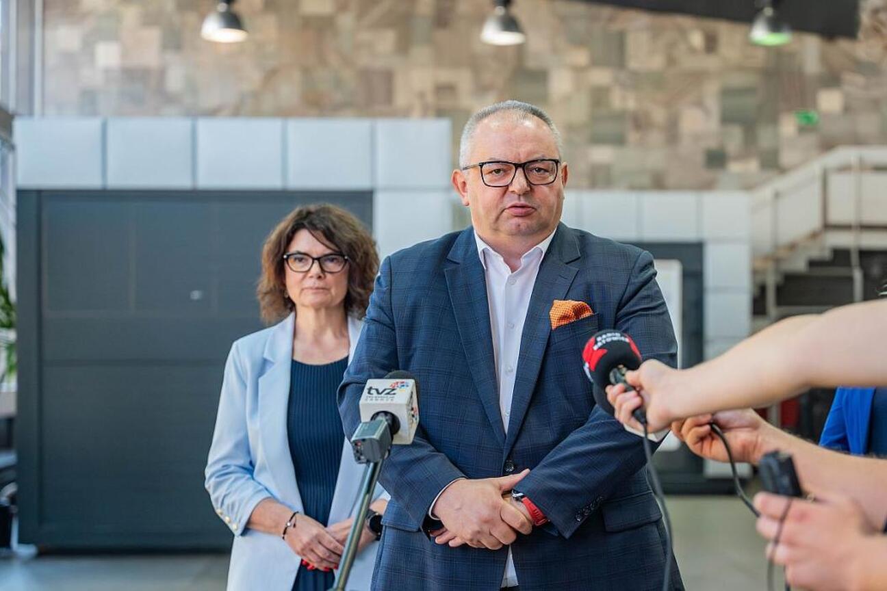 Mężczyzna w garniturze przemawia na konferencji prasowej, otoczony mikrofonami i dziennikarzami. W tle dwie kobiety, jedna z nich nosi okulary. Scena w nowoczesnym pomieszczeniu z kamienną ścianą i sc (wygenerowano przez AI)