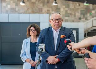 Mężczyzna w garniturze przemawia na konferencji prasowej, otoczony mikrofonami i dziennikarzami. W tle dwie kobiety, jedna z nich nosi okulary. Scena w nowoczesnym pomieszczeniu z kamienną ścianą i sc (wygenerowano przez AI)