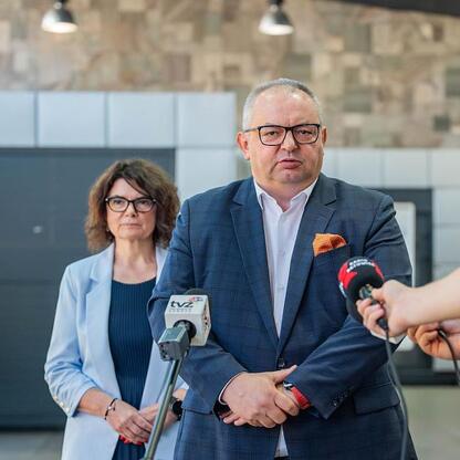 Mężczyzna w garniturze przemawia na konferencji prasowej, otoczony mikrofonami i dziennikarzami. W tle dwie kobiety, jedna z nich nosi okulary. Scena w nowoczesnym pomieszczeniu z kamienną ścianą i sc (wygenerowano przez AI)