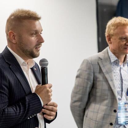 Podczas uroczystości wręczenia Nagród Marszałka Województwa Śląskiego na Superauto.pl Stadion Śląski, dwaj uczestnicy w czarnym i szarym garniturze uczestniczą w ceremonii, jednego z nich prowadzi mik (wygenerowano przez AI)