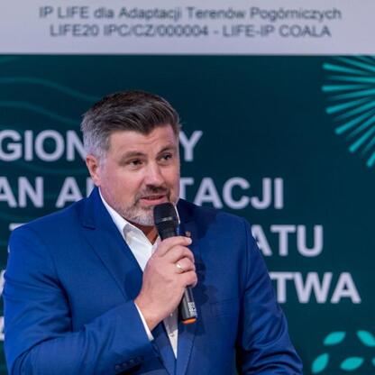 Mężczyzna w niebieskim garniturze przemawia do mikrofonu na konsultacjach społecznych w ramach Regionalnego Planu Adaptacji do zmian klimatu dla Województwa Śląskiego. (wygenerowano przez AI)