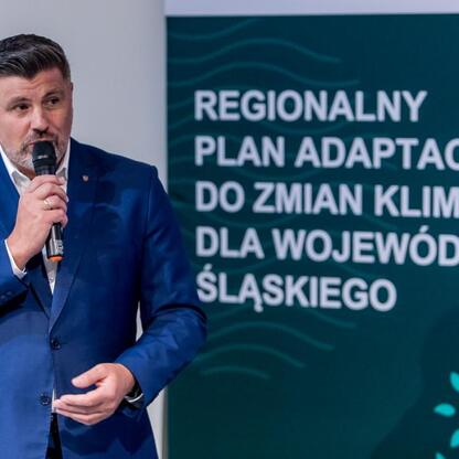 Mężczyzna w niebieskim garniturze przemawia do mikrofonu, prezentując Regionalny Plan Adaptacji do Zmian Klimatu dla Województwa Śląskiego. (wygenerowano przez AI)