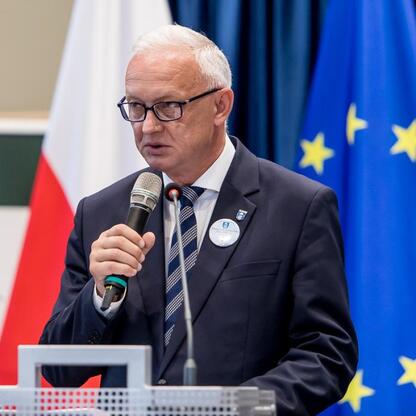 Mężczyzna w ciemnym garniturze przemawia do publiczności, tło: flagi UE i Polski, uroczystość inauguracji roku szkolnego 2025/26. (wygenerowano przez AI)