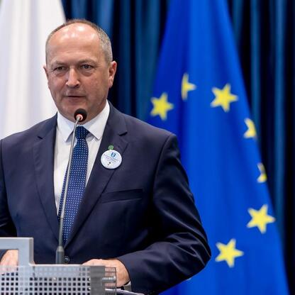 Mężczyzna w ciemnym garniturze i krawacie przemawia do mikrofonu, tło: flagi Polski i Unii Europejskiej, uroczystość w Kłobucku. (wygenerowano przez AI)