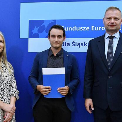 Członkowie Zarządu Województwa Śląskiego Rafał Adamczyk i Joanna Bojczuk wręczają listy gratulacyjne w ramach programu Fundusze Europejskie dla Śląskiego 2021-2027. (wygenerowano przez AI)