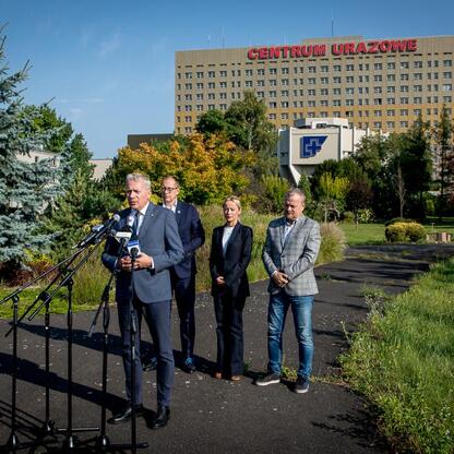 Grupa osób podczas konferencji prasowej przed Centrum Urzędowym, omawiając przejęcie terenu dla Wojewódzkiego Szpitala Specjalistycznego nr 5 im. św. Barbary w Sosnowcu. (wygenerowano przez AI)