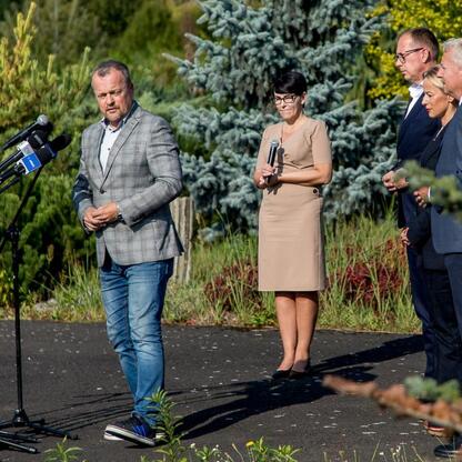 Grupa czterech osób w formalnym stroju przemawia do mikrofonów, podczas konferencji prasowej dotyczącej transferu terenu dla Wojewódzkiego Szpitala Specjalistycznego nr 5 im. św. Barbary w Sosnowcu. (wygenerowano przez AI)