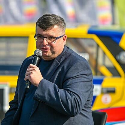 Na zdjęciu: Mężczyzna w ciemnym garniturze i okularach przesłuchuje się w mikrofonie podczas konferencji prasowej Rajdu Śląska. W tle widoczny jest żółty samochód z numerem 