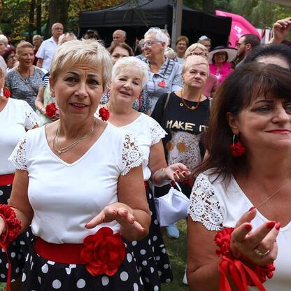 Grupa kobiet w białych bluzach i spódnicach w kwiaty tańczy na Silver Silesia, festiwalu dla seniorów. (wygenerowano przez AI)