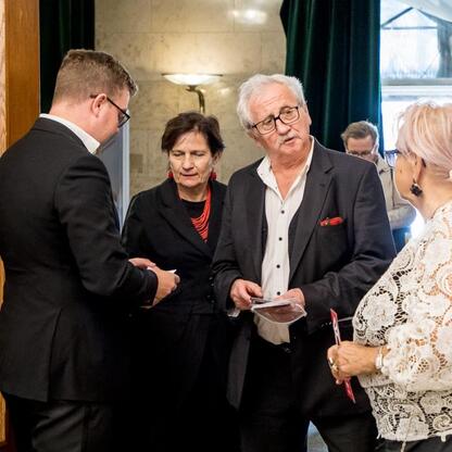 Grupa ludzi w formalnym stroju rozmawia w sali konferencyjnej, związana z inicjatywą 