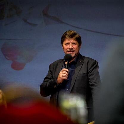 Na scenie Teatru Śląskiego mąż w ciemnym płaszczu przemawia do publiczności, tło abstrakcyjne. (wygenerowano przez AI)