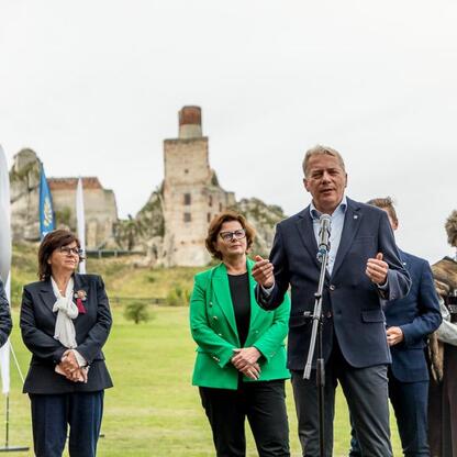 Zapowiedź konferencji prasowej Juromania, która odbędzie się w dniach 19-21 września. Marszałek Wojciech Saługa i inne dostojnicy uczestniczą w uroczystości na tle ruin Olsztynu. (wygenerowano przez AI)