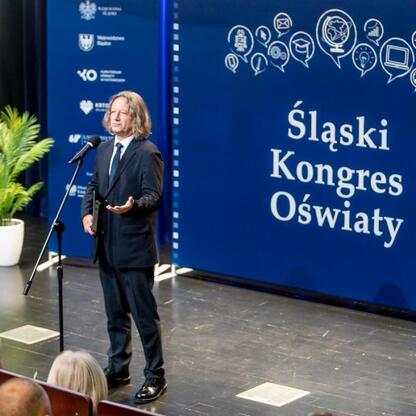 Na Śląskim Kongresie Oświaty w Katowicach, na scenie stoi mówca w garniturze, prezentujący wyzwanie dotyczące edukacji. (wygenerowano przez AI)