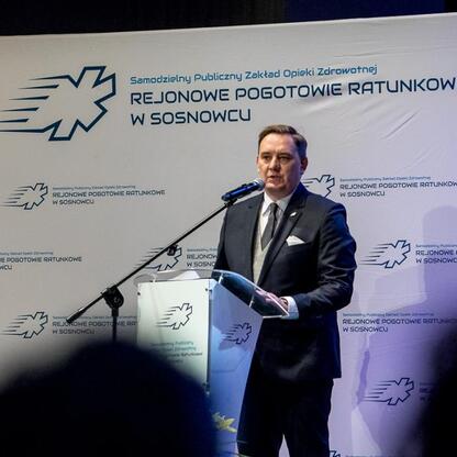 Mężczyzna w garniturze przemawia na podium podczas uroczystości jubileuszowej 30-lecia SPZOZ Rejonowego Pogotowia Ratunkowego w Sosnowcu. (wygenerowano przez AI)