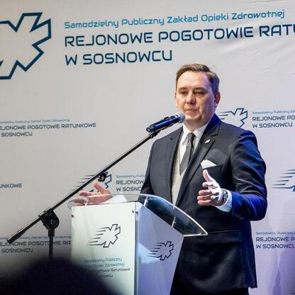 Mężczyzna w ciemnym garniturze przemawia na podium podczas uroczystości 30-lecia Sosnowieckiego Pogotowia Ratunkowego. (wygenerowano przez AI)