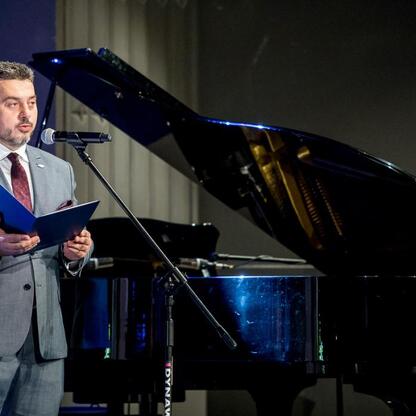 Uroczystość 30-lecia Sosnowieckiego Pogotowia Ratunkowego w Pałacu Kultury Zagłębia. Mężczyzna w garniturze przemawia z mikrofonem, trzymając folder. (wygenerowano przez AI)