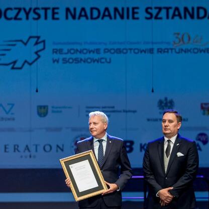 Marszałek Wojciech Saługa i inny wojewódzki urzędnik uczestniczą w uroczystości 30-lecia Sosnowieckiego Pogotowia Ratunkowego. (wygenerowano przez AI)