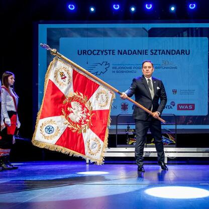 Marszałek Wojciech Saługa prezentuje sztandar na uroczystości 30-lecia Sosnowieckiego Pogotowia Ratunkowego. (wygenerowano przez AI)