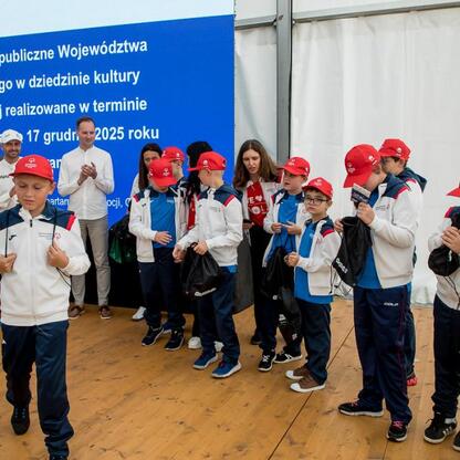 Grupa młodych sportowców w czerwonych czapkach i białych kurtkach stoi przed plakatem 
