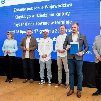 Grupa osób w strojach formalnych stoi przed niebieskim banerem z żółtym napisem 