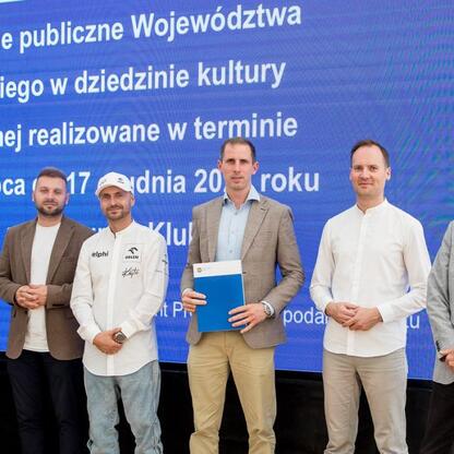 Grupa siedmiu osób w formalnym stroju stoi przed dużym niebieskim ekranem z tekstem w języku polskim. Wydarzenie związane z 