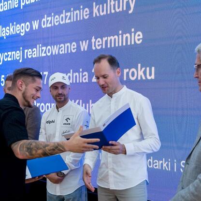 Formalna uroczystość na Superauto.pl Stadionie Śląskim, gdzie Leszek Pietraszek i Kajetan Kajetanowicz wręczają symboliczne czeki przedstawicielom 154 klubów sportowych z regionu. (wygenerowano przez AI)