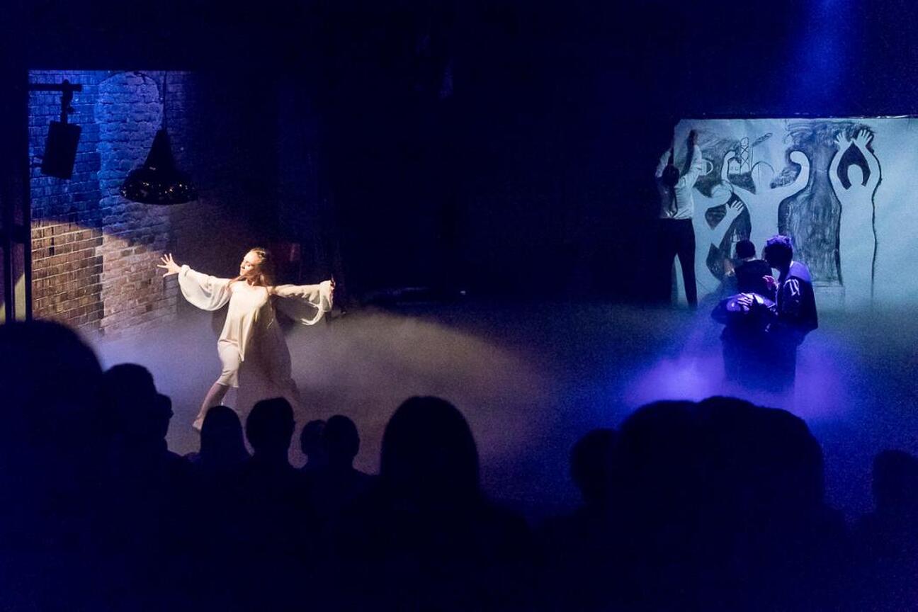 Teatralna scena z aktorami w ekspresyjnych ruchach pod dramatycznym oświetleniem. W tle ceglane ściany i abstrakcyjna kompozycja, widoczna silueta widzów. (wygenerowano przez AI)