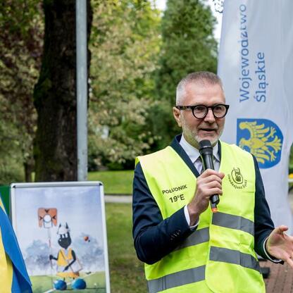 Mężczyzna w żółtym kamizelce z logo Województwa Śląskiego, trzymający mikrofon, stoi przed flagami i banerem z napisem 