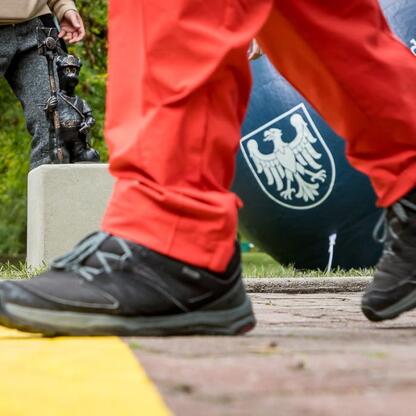 Zdjęcie przedstawia zbliżenie na nogi i buty osoby, która kroczem przekroczyła żółtą linię w Katowickim Parku Kościuszki, z figurką gnomu i sztandarem z orłem na tle. (wygenerowano przez AI)