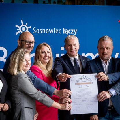 Grupa siedmiu osób w strojach formalnych, trzymających dokument, przedstawiająca projekt rewitalizacji terenu byłej kopalni KWK Kazimierz-Juliusz w Sosnowcu. (wygenerowano przez AI)