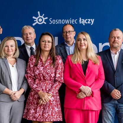 Grupa oficjalnych osób w koszulach z logo Śląska, przedstawiająca projekt rewitalizacji byłego KWK Kazimierz-Juliusz w Sosnowcu. (wygenerowano przez AI)