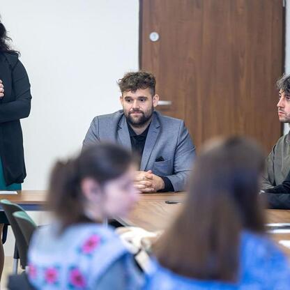 Formalna sesja w konferencyjnym pomieszczeniu, gdzie przedstawicielka z Ukrainy w czarnym płaszczu i zielonej sukience przemawia do grupy uczestników w strojach biznesowych, siedzących wokół długiego  (wygenerowano przez AI)