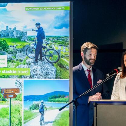 Podczas Wojewódzkich Obchodów Światowego Dnia Turystyki w Tarnowskich Górach, organizowanych przez Tarnogórskie Centrum Kultury, na podium stoją dwaj goście honorowi, którzy dostarczają informacje o r (wygenerowano przez AI)