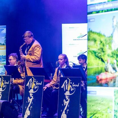 Występują na zdjęciu muzycy grający na saksofonach i innych instrumentach, podczas koncertu jazzowego na scenie podczas Wojewódzkich Obchodów Światowego Dnia Turystyki w Tarnowskich Górach. (wygenerowano przez AI)