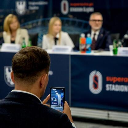 Na zdjęciu przedstawiono konferencję prasową z okazji 17. edycji Silesia Marathon, która odbywa się na Superauto.pl Stadion Śląski. Uczestnicy zapisują wydarzenie na smartfony. (wygenerowano przez AI)