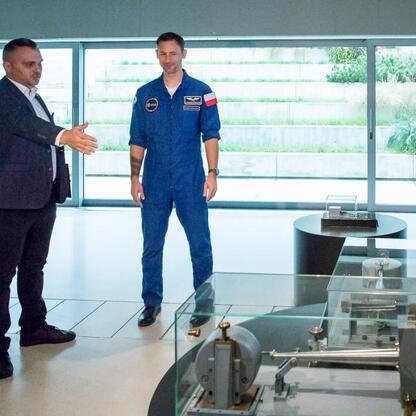 Dwoje osób w modernistycznym pawilonie nauki, jedna w garniturze, druga w kosmicznej odzieży NASA, dyskutują o kosmicznych wydarzeniach. (wygenerowano przez AI)