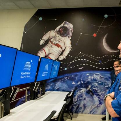 Astronauta w niebieskim strój kosmiczny prezentuje logo Śląskiego Parku Nauki na ekranie. (wygenerowano przez AI)